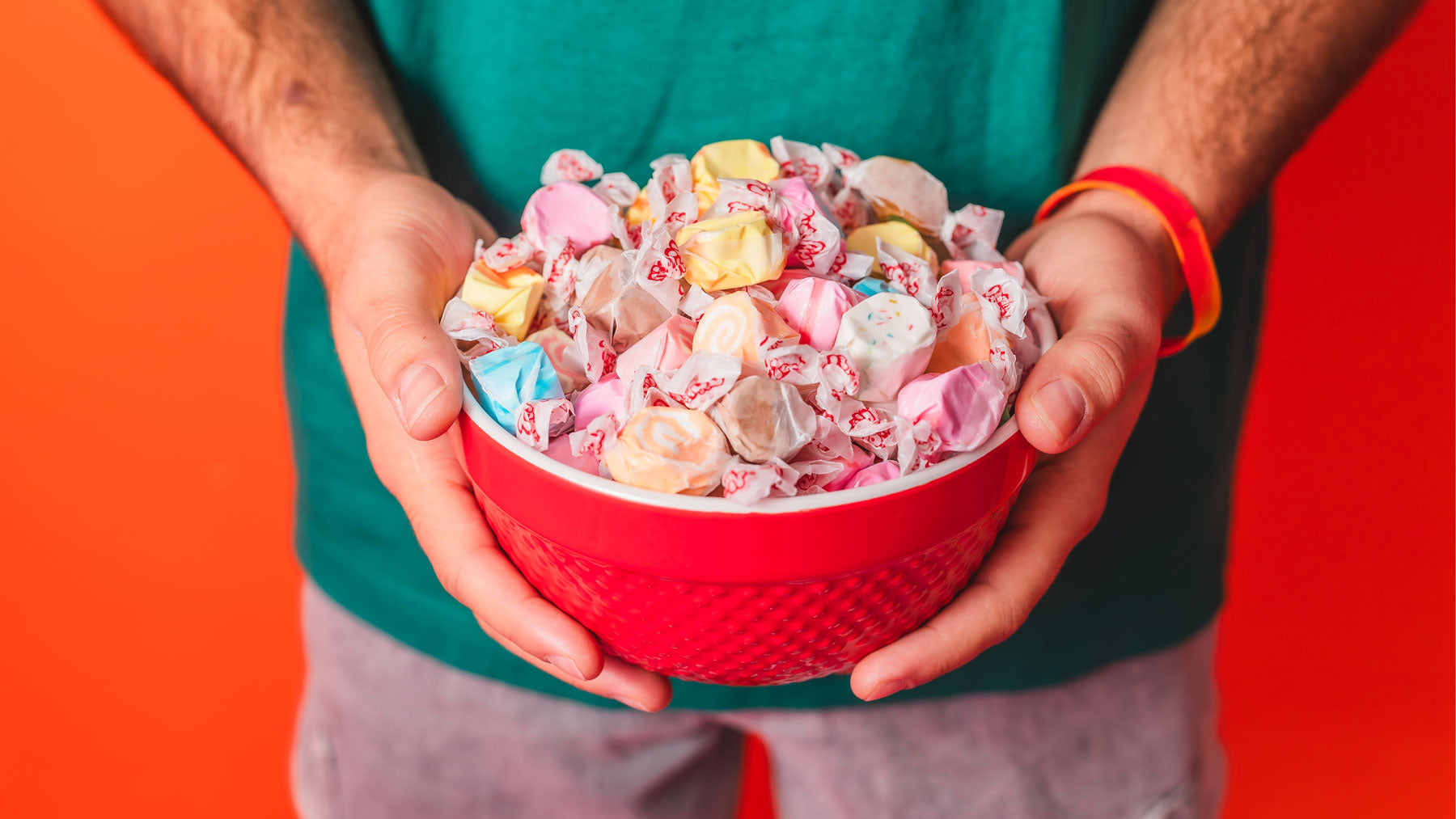 Hands holding a red bowl filled with assorted Taffy Town salt water taffy in pastels and brights, perfect for a party candy table.