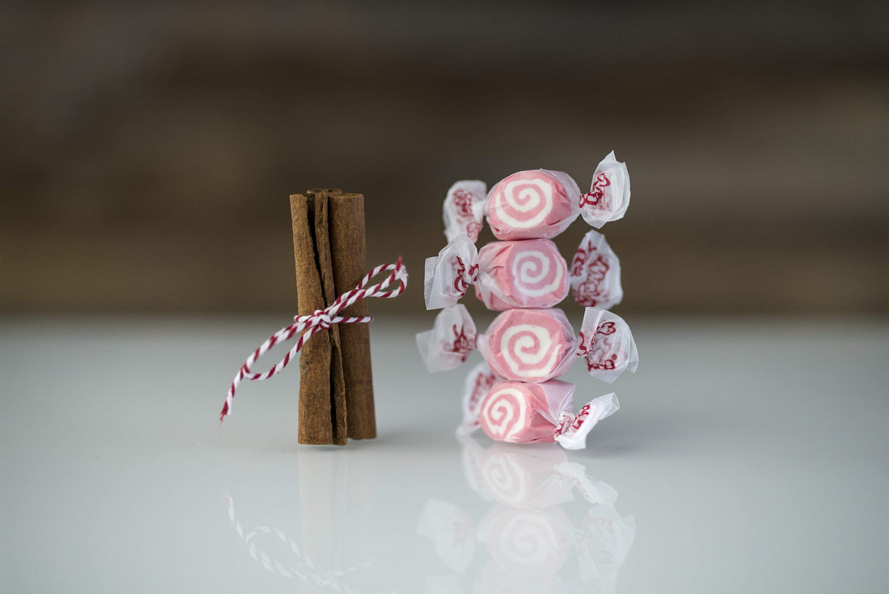 Five pieces of pink-and-white Cinnamon Swirls taffy candy from Taffy Town stacked beside tied cinnamon sticks—gourmet salt water taffy with a bold, spicy flavor.